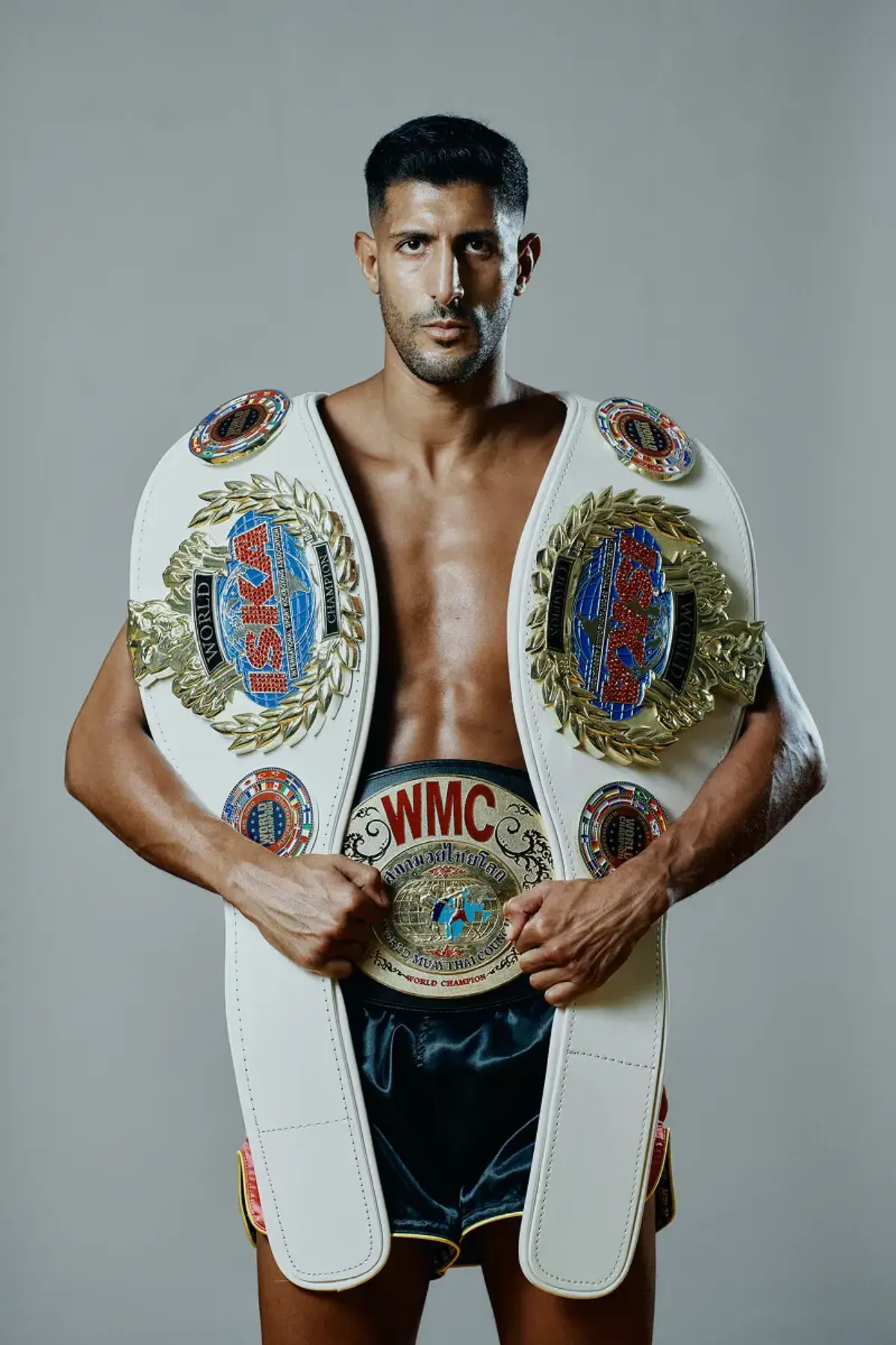 Youssef Assouik in a studio portrait, two ISKA world title belts over his shoulders and the WMC world title held at his waist