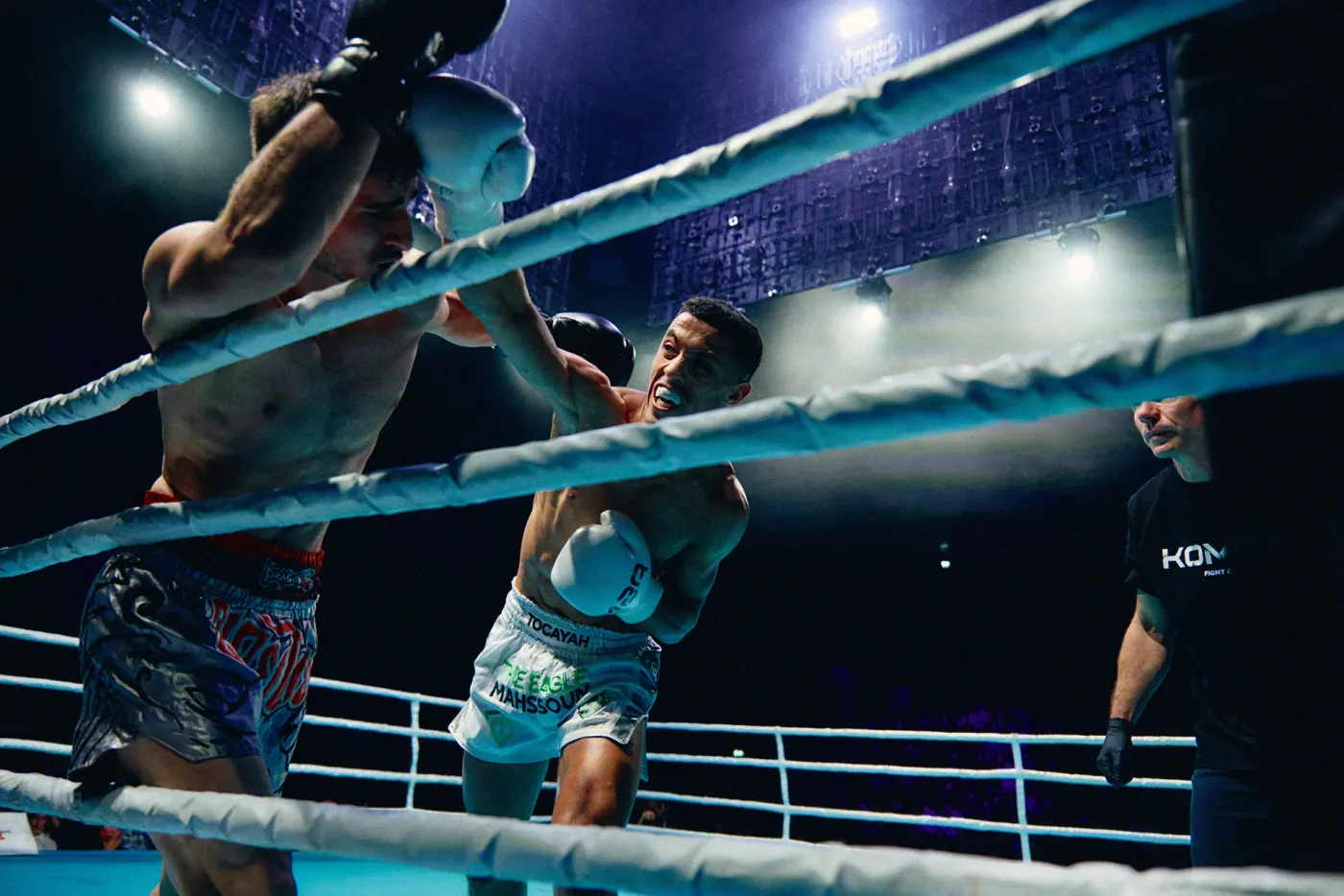 Two fighters exchanging punches on the ropes at K.B. Hallen, the referee a step away